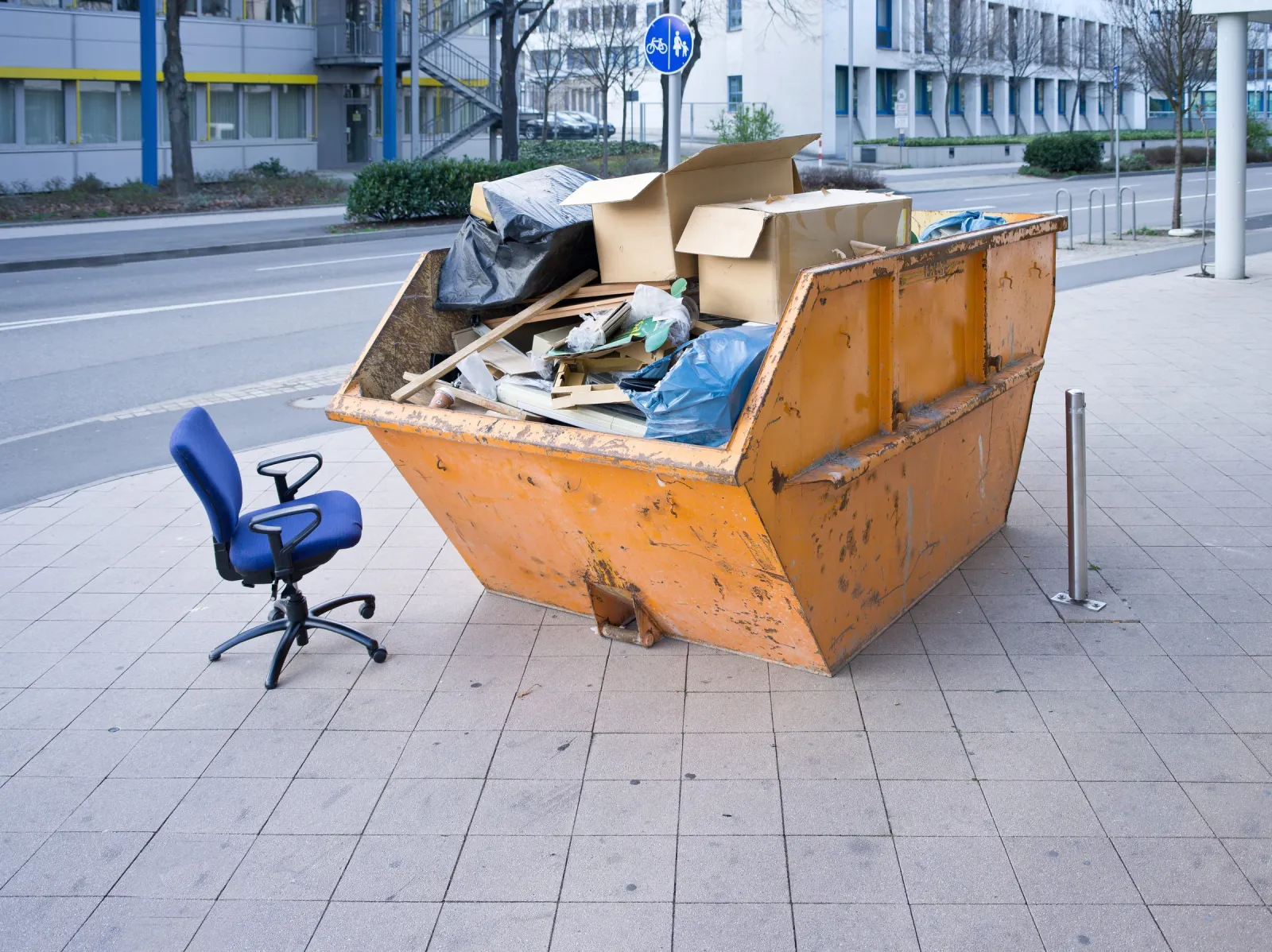 Absetzcontainer mit Möbeln und Kartons am Straßenrand, daneben ein Bürostuhl